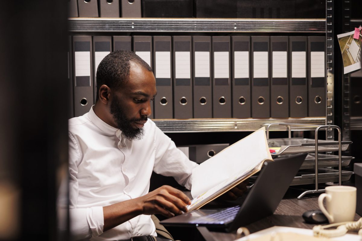 African american prosecutor reading file, preparing for case in police office with archival records. Law enforcement professional analyzing forensic expertise folder at night time